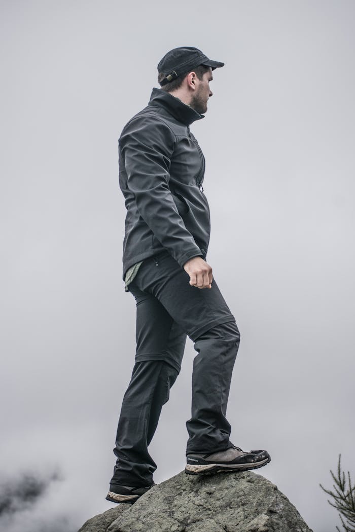 A man standing on a rock wearing a black jacket, looking over a cloudy landscape.